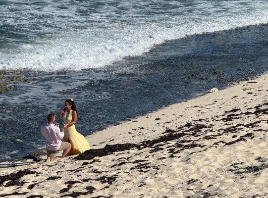 Ted proposes to Katy on a quiet Bahamas beach, creating a romantic moment with the ocean waves behind them.